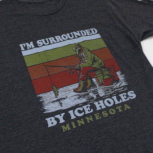 Dark gray t-shirt with graphic design of a man ice fishing and text that reads "I'm surrounded by ice holes" and "Minnesota" on a white background